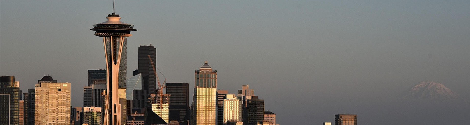 Im warmen Licht der untergehenden Sonne ist die Space Needle vor der beeindruckenden Skyline Seattles zu sehen, während Mount Rainier in der Ferne eine eindrucksvolle Verbindung von Stadtbild und Natur schafft.